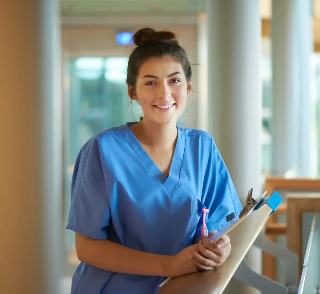 Enfermera sonriente con uniforme azul sosteniendo un portapapeles en un pasillo hospitalario luminoso.