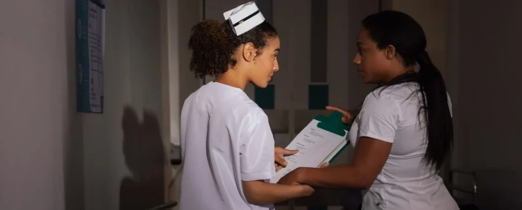 Dos enfermeras conversando en el pasillo de un hospital durante un turno, una revisa documentos clínicos con la otra.
