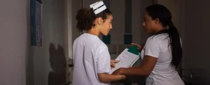 Dos enfermeras conversando en el pasillo de un hospital durante un turno, una revisa documentos clínicos con la otra.