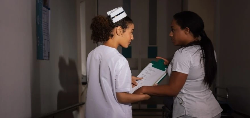 Dos enfermeras conversando en el pasillo de un hospital durante un turno, una revisa documentos clínicos con la otra.